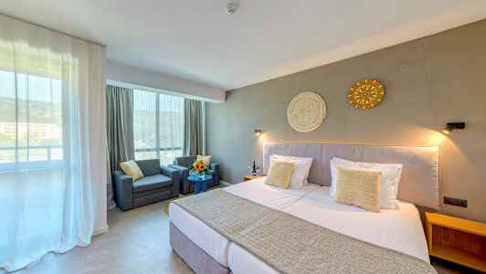 Hotellihuone, jossa on suuri sänky, kaksi nojatuolia pöydän ääressä ja koriste seinällä. (Tekoälyn luoma teksti)