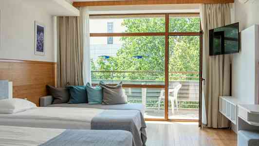 Hotellihuone, jossa on kaksi sänkyä, sohva värikkäillä tyynyillä ja lasiovi, joka johtaa parvekkeelle. (Tekoälyn luoma teksti)