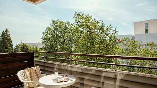 Parveke, jossa pöytä, kaksi muovituolia, laukku, aurinkolasit ja sanomalehti, näkymä puihin ja rakennuksiin. (Tekoälyn luoma teksti)