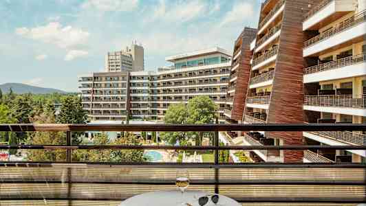 Näkymä parvekkeelta, jossa on pöytä ja lasi, hotellirakennuksille ja uima-allasalueelle. (Tekoälyn luoma teksti)