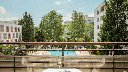 Näkymä parvekkeelta, jonka eteen avautuu uima-allas ja vehreää ympäristöä; pöydällä lasi viiniä ja kirja. (Tekoälyn luoma teksti)