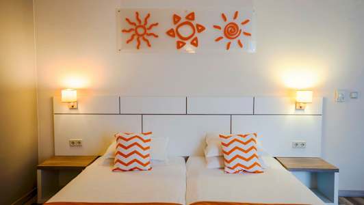 Hotellihuone, jossa on kaksi erillistä vuodetta, oranssit tyynyt ja seinäkoristeita aurinkojen muodossa. (Tekoälyn luoma teksti)