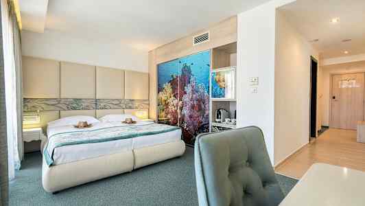 Kuva hotellihuoneesta, jossa on parivuode, merihenkinen sisustus sängyn takana ja työpöytä tuoleineen. (Tekoälyn luoma teksti)