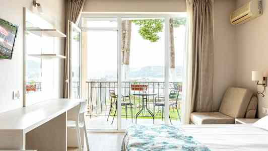 Hotellihuone, jossa on sänky, pöytä ja tuoli, sekä parveke ulkokalusteineen ja näkymä luontoon. (Tekoälyn luoma teksti)