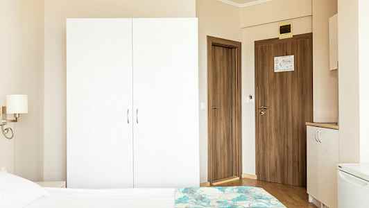 Hotellihuoneen ympäristö, jossa valkoinen vaatekaappi, puuovia ja osa sängystä etualalla. (Tekoälyn luoma teksti)