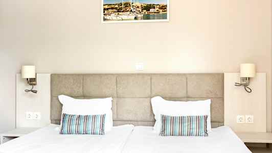 Kaksi sänkyä, joissa valkoiset vuodevaatteet ja raidalliset tyynyt, seinällä taulu ja yöpöytälamput. (Tekoälyn luoma teksti)