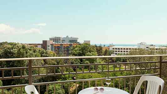 Näkymä parvekkeelta, jossa on pöytä ja tuoli, taustalla vihreää aluetta, rakennuksia ja meri. (Tekoälyn luoma teksti)