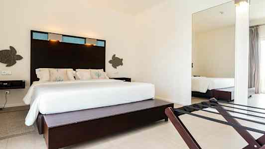 Hotellihuone, jossa on suuri sänky valkoisilla vuodevaatteilla ja koristeellisilla tyynyillä sekä tummat puupaneelit sängyn takana. (Tekoälyn luoma teksti)