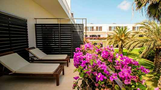 Terassi, jossa kaksi aurinkotuolia ja kukkivia kasveja, näkymä puutarhaan ja rakennuksiin. (Tekoälyn luoma teksti)