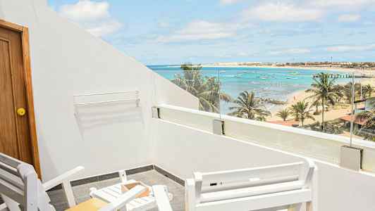 Kuvassa on ulkoterassi, jolla on valkoisia tuoleja ja pöytiä sekä näkymä rannalle, palmuihin, mereen ja pieniin veneisiin. (Tekoälyn luoma teksti)