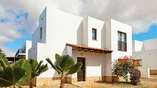 Valkoinen rakennus, jossa on kivipohja, ympärillä kasveja ja puinen sisäänkäynti. (Tekoälyn luoma teksti)