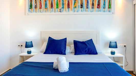 Kuvassa on sänky, jossa on sinisiä tyynyjä ja vuodevaatteita, vierellään kaksi yöpöytää, joissa siniset lamput. (Tekoälyn luoma teksti)