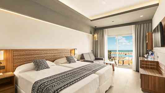 Hotellihuone, jossa on kaksi sänkyä ja oleskelutila, lasioven vieressä, joka johtaa parvekkeelle näkymällä. (Tekoälyn luoma teksti)