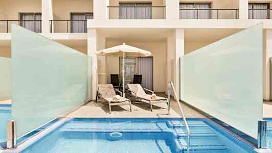 Allasalue, jossa on kaksi aurinkotuolia ja aurinkovarjo valkoisen parvekkeellisen rakennuksen edessä. (Tekoälyn luoma teksti)