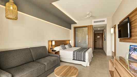 Nykyaikainen hotellihuone, jossa on sänky, sohva ja pöytä. Seinällä on myös litteänäyttöinen televisio. (Tekoälyn luoma teksti)