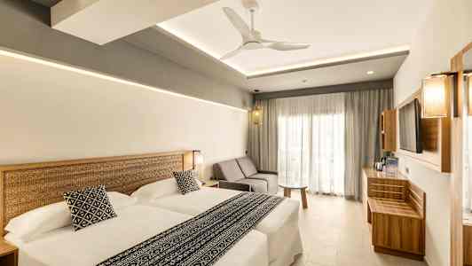 Moderni hotellihuone, jossa on kaksi sänkyä, sohva ja pöytä. Tyyli on valoisa ja minimalistinen. (Tekoälyn luoma teksti)