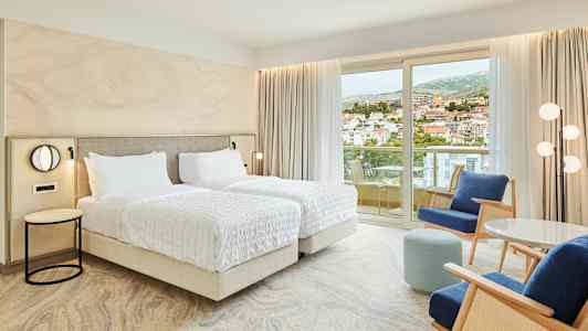 Hotellihuone, jossa on kaksi yhden hengen vuodetta, istuma-alue ja parveke näkymällä kaupunkiin. (Tekoälyn luoma teksti)