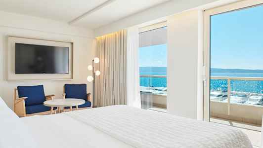 Hotellihuoneen sisätilat, joissa on suuri sänky, kaksi tuolia, pöytä ja televisio. Näkymä merelle parvekkeen oven kautta. (Tekoälyn luoma teksti)