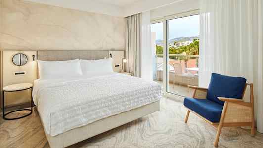 Makuuhuone, jossa suuri sänky ja sininen tuoli lasiovien lähellä parvekkeelle. (Tekoälyn luoma teksti)