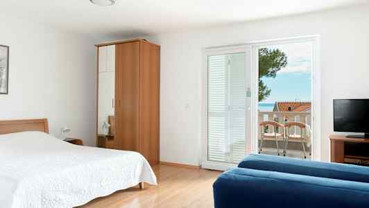 Hotellihuone, jossa on sänky, vaatekaappi, sininen sohva, televisio ja parveke näkymällä. (Tekoälyn luoma teksti)