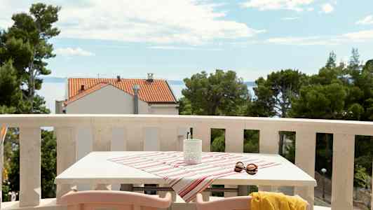 Parveke, jossa pöytä peitetty raidallisella liinalla, aurinkolasit ja keltainen huivi, näkymä merelle ja taloja. (Tekoälyn luoma teksti)