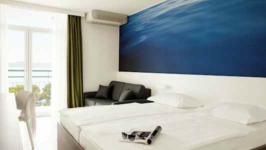 Kahden hengen huone, jossa suuri sininen seinämaalaus sängyn takana. Parveke merinäköalalla. (Tekoälyn luoma teksti)