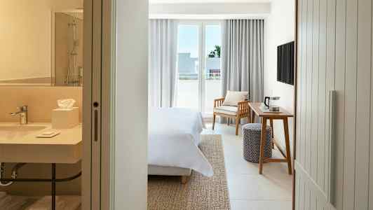 Hotellihuone, jossa on sänky, kirjoituspöytä, tuoli ja parveke. Etualalla näkyy kylpyhuone. (Tekoälyn luoma teksti)