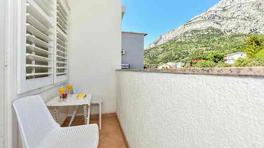 Parveke, jolla on kaksi valkoista tuolia, pöytä, jossa on kaksi lasia mehua ja hedelmiä, sekä näkymä vuorille ja ympäröiviin rakennuksiin. (Tekoälyn luoma teksti)