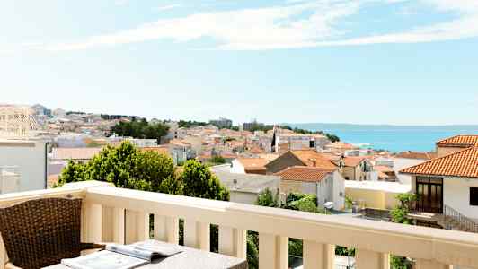 Parveke, jossa pöytä ja tuoli, näkymä kaupungille, jossa punaisia kattoja, ja taustalla meri. (Tekoälyn luoma teksti)