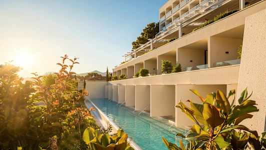 Hotellirakennus, jonka edessä pitkä uima-allas, ympäröitynä kasveilla ja auringonnousulla. (Tekoälyn luoma teksti)