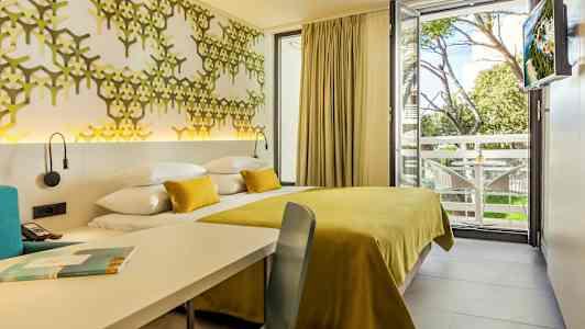 Valokuva hotellihuoneesta, jossa on keltainen sängynpäällinen ja koristeellinen seinäpaneeli sekä näkymä puistoalueelle ja parvekkeelle. (Tekoälyn luoma teksti)