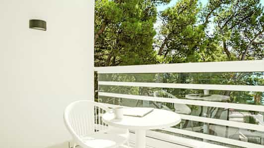 Parveke, jossa valkoinen pyöreä pöytä ja valkoinen tuoli. Näkymä vihreisiin puihin. (Tekoälyn luoma teksti)