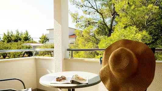 Parveke, jossa pöytä, tuoli ja aurinkohattu, näkymä puille ja rakennuksille. (Tekoälyn luoma teksti)