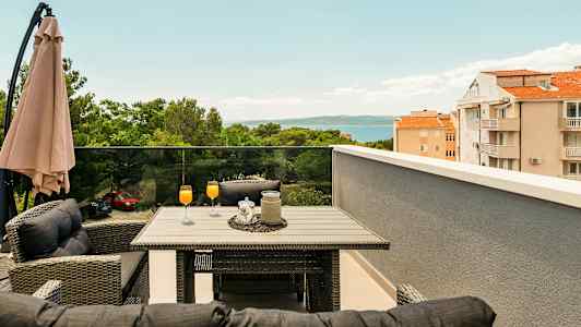 Terassi, jossa pöytä ja tuolit, oransseja juomia laseissa, varjo sekä näkymä puihin, rakennuksiin ja merelle. (Tekoälyn luoma teksti)