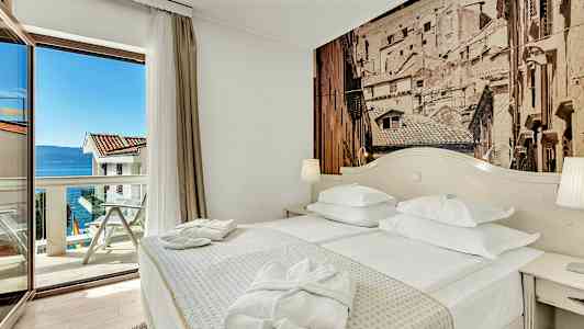 Hotellihuone, jossa on parivuode, pöytä ja lasiovi, joka johtaa parvekkeelle merinäköalalla. (Tekoälyn luoma teksti)