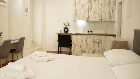 Kuva vaaleasti sisustetusta huoneesta, jossa on sänky, keittiötaso ja pöytä tuoleineen. (Tekoälyn luoma teksti)