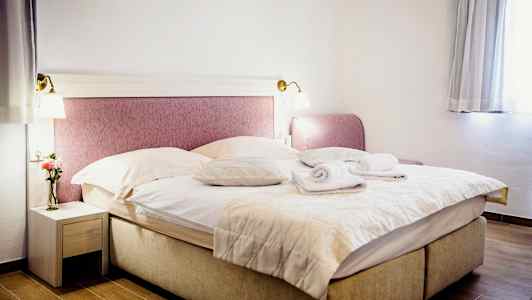 Huone, jossa on parivuode, pedattu peitoilla ja tyynyillä, päällä kaksi rullattua pyyhettä. Yöpöytä vasemmalla maljakolla ja kukalla sekä ikkuna oikealla verhoineen. (Tekoälyn luoma teksti)