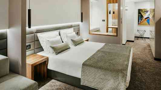 Moderni hotellihuone, jossa on suuri sänky, valkoinen vuodevaatteet, harmaa päiväpeite ja tyynyt sekä harmaa kokolattiamatto. (Tekoälyn luoma teksti)