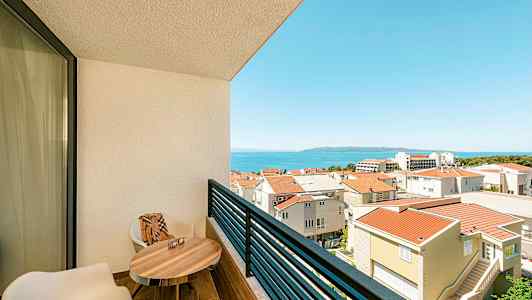 Näkymä parvekkeelta, jolla on kaksi tuolia, pöytä ja näkymä kaupunkialueelle ja merelle. (Tekoälyn luoma teksti)