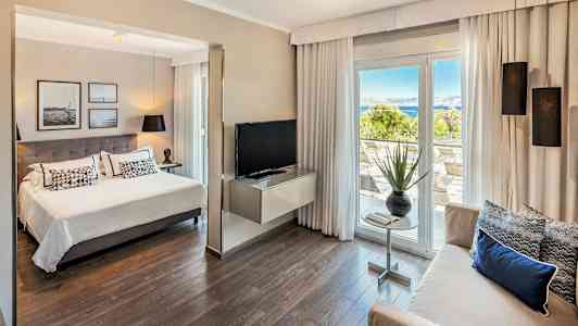 Hotellihuoneen sisätila, jossa istuinryhmä, sänky ja näkymä terassille sekä taustalla meri. (Tekoälyn luoma teksti)
