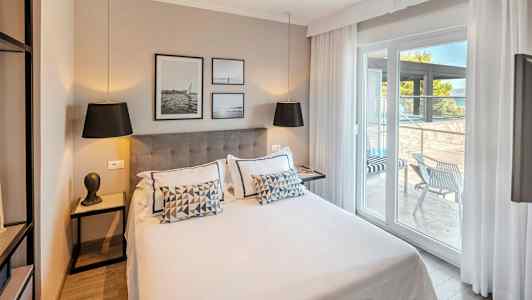 Moderni hotellihuone, jossa on parivuode, koristeltu tyynyillä ja taiteella seinällä. Lasiovet johtavat sisustetulle terassille. (Tekoälyn luoma teksti)