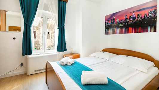 Makuuhuone, jossa on parivuode, turkoosit verhot ja seinällä kuva kaupungin siluetista. (Tekoälyn luoma teksti)