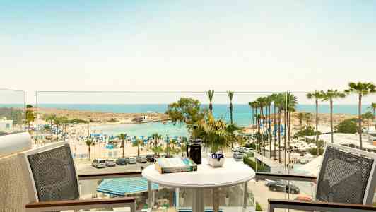 Näkymä parvekkeelta, jossa pöytä ja tuolit, rannalle, jolla on aurinkovarjoja ja palmuja meren äärellä. (Tekoälyn luoma teksti)