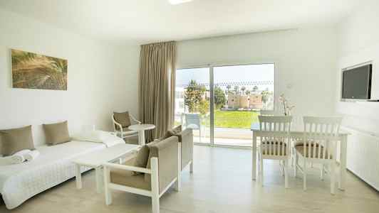 Vaaleasti sisustettu huone, jossa sänky, tuolit, pöytä ja näkymä viheralueelle suurten ikkunoiden kautta. (Tekoälyn luoma teksti)