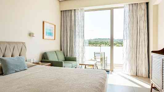 Hotellihuone, jossa on sänky, sohva ja pöytä, sekä parveke tuoleineen ja näkymä vehreyteen. (Tekoälyn luoma teksti)