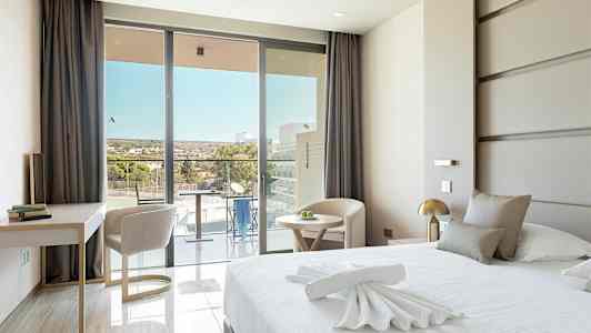 Moderni hotellihuone, jossa on suuri sänky, kirjoituspöytä ja tuoleja sekä parveke näkymällä. (Tekoälyn luoma teksti)