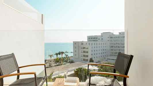 Kaksi tuolia ja pöytä parvekkeella näkymällä hotellirakennuksiin, palmuihin ja mereen. (Tekoälyn luoma teksti)