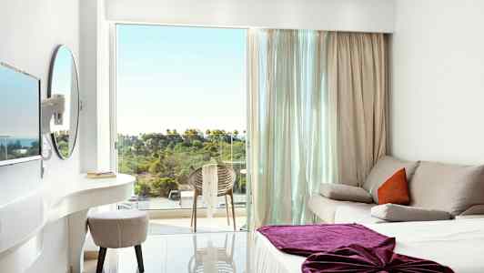 Vaalea hotellihuone, jossa sohva, sänky violetilla peitteellä ja parveke, jolta näkyy vehreää maisemaa. (Tekoälyn luoma teksti)