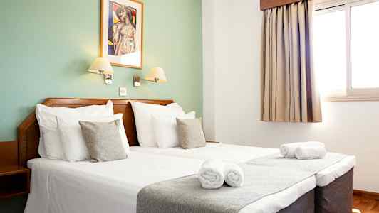 Hotellihuoneen sisätilat, jossa parivuode, kaksi sängyn lamppua, ikkuna verhoilla ja seinäkoriste. (Tekoälyn luoma teksti)