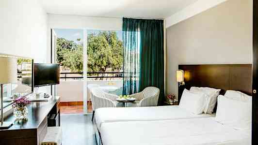 Hotellihuone, jossa on kaksi sänkyä, kirjoituspöytä ja pieni pöytä tuoleineen lasioven edessä. (Tekoälyn luoma teksti)